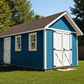 Blue storage shed with white doors on a grassy area with trees in the background