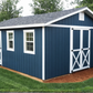 Blue storage shed with white trim on a grassy area with trees in the background